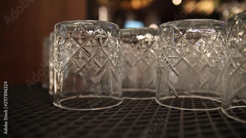 Empty inverted crystal cut-glass tumbler glasses resting on a black rubber bar mat surface with blurred warm bokeh lights and a cozy nightlife pub restaurant interior atmosphere.