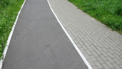 Green urban park pathway combining asphalt walking trail and paved sidewalk surrounded by grass and natural landscape