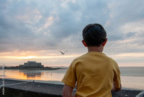 Fotografie Au coucher du soleil, vu de dos, un jeune enfant au pull jaune contemple la mer