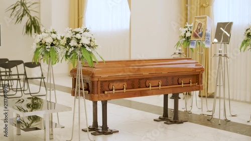 Closed wooden coffin in a bright funeral home or chapel. Solemn farewell ceremony setup with white floral arrangements, empty chairs, and a religious icon.