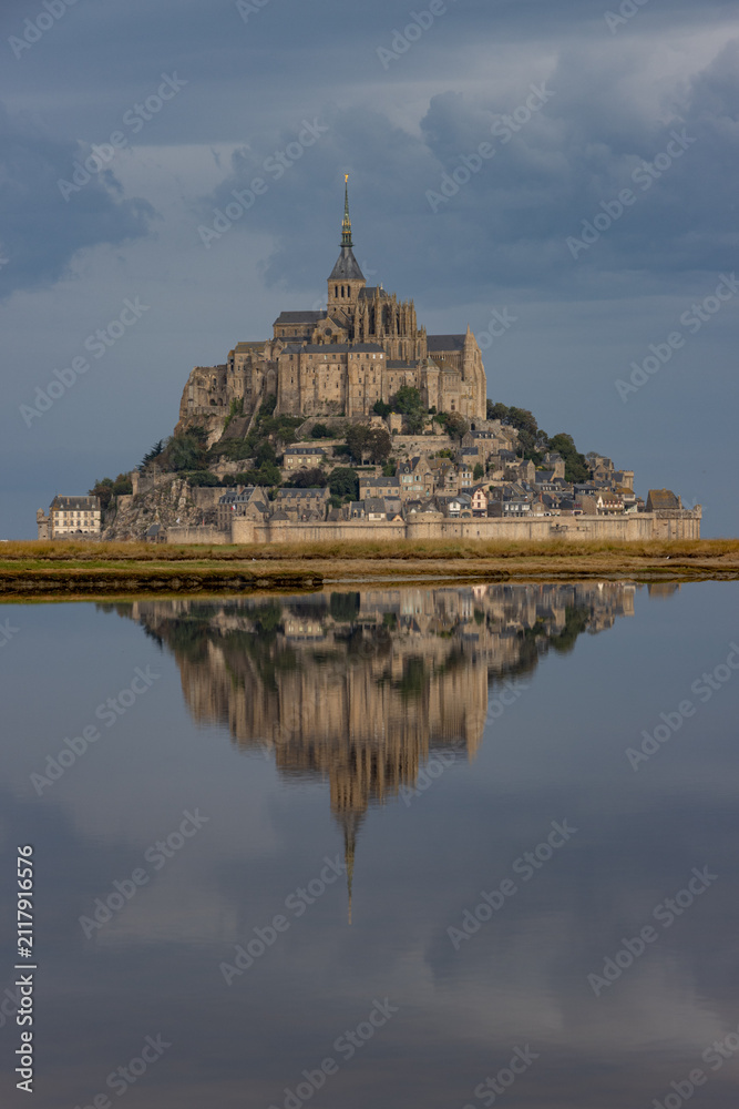 Fototapeta premium Atmospheric vertical shot of Mont Saint-Michel casting a pristine mirror reflection onto calm tidal waters under a dark stormy sky in Normandy.