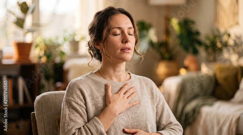 Serene Respite: A woman sits with eyes closed, hand on her chest, deeply inhaling. Sunlight streams into a cozy, plant-filled room, highlighting a moment of peaceful meditation and self-care.