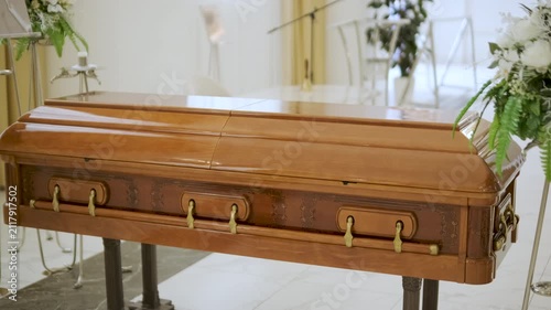 Closed wooden coffin in a bright funeral home or chapel. Solemn farewell ceremony setup with white floral arrangements, empty chairs, and a religious icon.