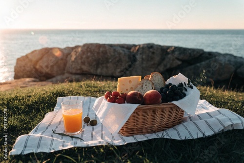 Wicker basket with tasty food (cheese, bread and fruits) and juice for romantic picnic near sea at sunset 