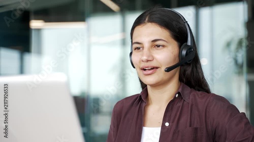 Close up of female customer service operator in headset working on laptop and talking to client in modern office. Woman support agent provides assistance, answers questions and helps solve issues.