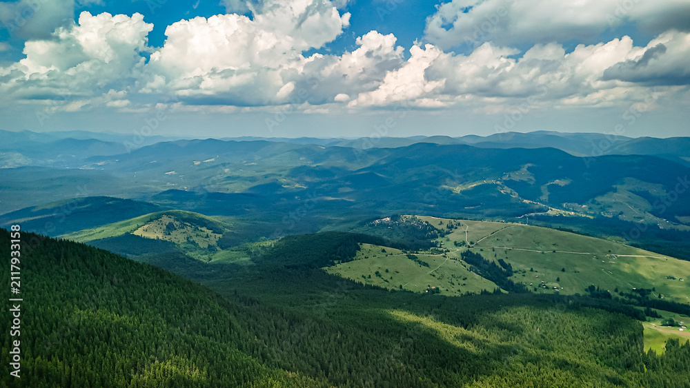 Fototapeta premium Rolling ranges drift beneath a bright sky as forests and meadows share the slopes. The aerial scene captures distance depth and light that shifts across a timeless mountain realm.