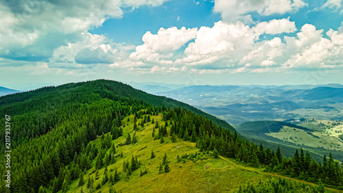 From a breezy crest the landscape opens to valleys glimmering through haze. Emerald ridges carry scattered pines while playful clouds cast roaming shadows across remote countryside