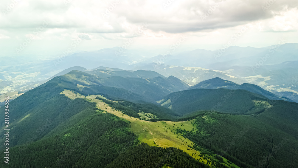 Fototapeta premium Broad ridges of conifer forest tilt toward a glimmering lowland. Patches of sunshine move across the slopes while the far ranges melt into haze beneath a roof of weighty clouds