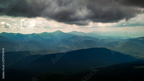 Green ridges fold like waves around quiet basins while pale light filters through cloud. The aerial view reveals serene forests open saddles and distant towns set in wide mountain realm