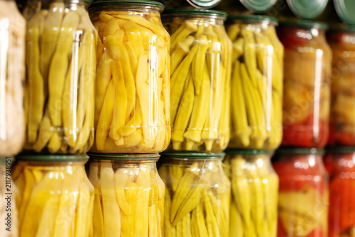Assorted canned vegetables, okra, yellow beans, and red bell peppers arranged in glass jars on shelf. Preserves prepared for wholesale trade and grocery distribution.