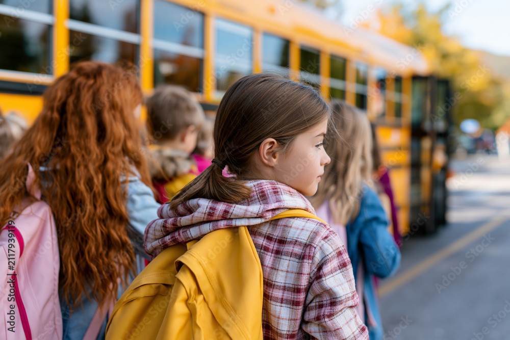 Obraz premium Young girl waits with classmates near school bus. She wears plaid jacket and carries yellow backpack. Her hair is tied back neatly, gaze focused ahead. Children stand in quiet line, anticipating ride