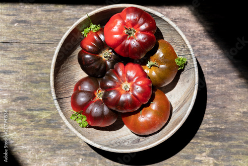 Organic heirloom tomato variety in wooden bowl with parsley on rustic wood table, harsh sunlight and deep shadow, flat lay