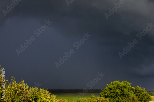 Foto Massive storm clouds loom over bright green fields while sunlight brushes the tr