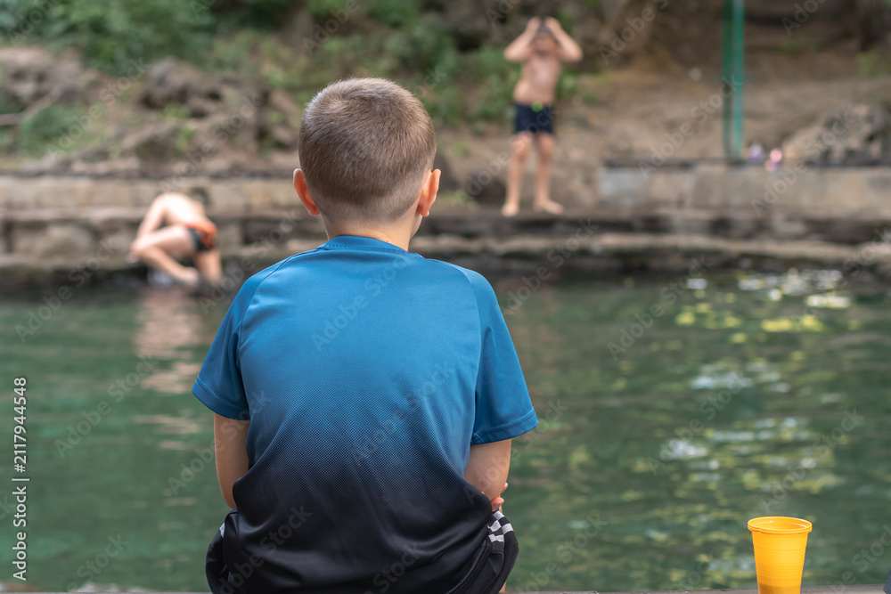 Fototapeta premium A boy sitting on the edge of a pond and watching other children swimming.