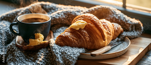 Croissant with butter and coffee cup on a wooden surface warm lighting breakfast pastry viennoiserie beverage hot drink caffeine spread tabletop still life