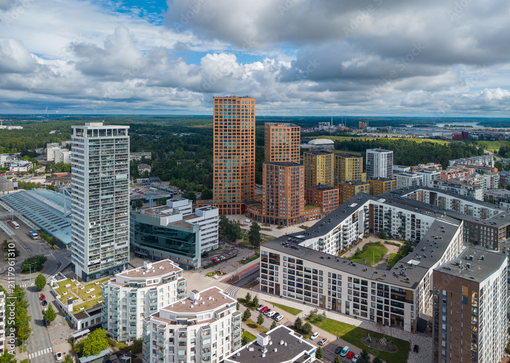 Fototapeta premium Aerial view of Vuosaari, Helsinki, showcasing a mixed skyline with tall glass tower, orange-brick high-rises, residential blocks, surrounding forest, and distant harbor under cloudy sky