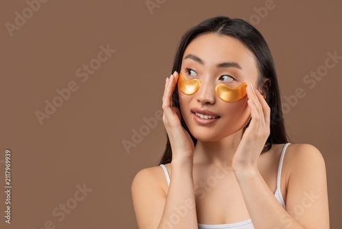 Smiling Young Asian Woman Applying Golden Eye Patches On Her Face In Front Of Mirror At Home, Taking Care Of Skin With A Skincare Routine. Cozy Home Setting, Wellness Concept.