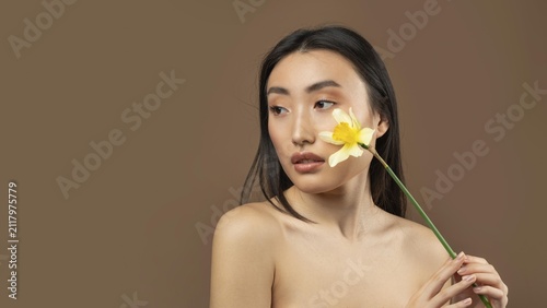Fotografie Thoughtful Young Asian Woman Holding Flower Near Her Face, Looking Away, In Studio Setting, Displaying Natural Beauty And Serenity