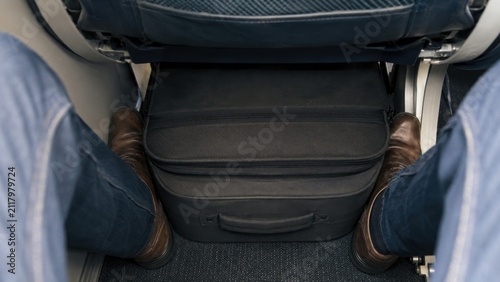 Passenger's legs in blue jeans positioned beside a black carry-on bag under an airplane seat, showcasing limited legroom and discomfort during flight