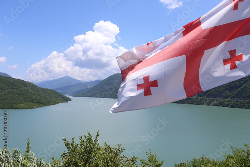 National flag of Georgia waving over turquoise water of Zhinvali Reservoir and Caucasian mountains landscape