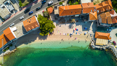 Фотография Aerial top view of people sunbathing on a sandy beach with turquoise water and orange-roofed houses in a Mediterranean coastal town