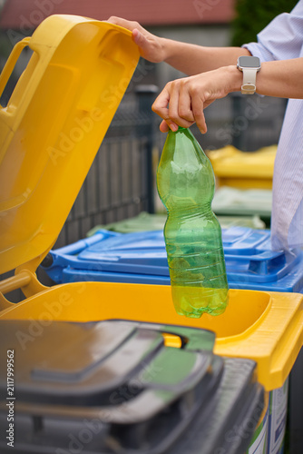 Hand placing empty plastic bottle into yellow recycling bin outdoors