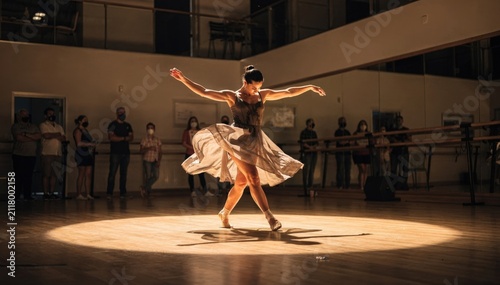 Medium shot of a dance academy open house spotlight on a lead dancer performing a fluid routine other performers and audience softly out of focus behind.