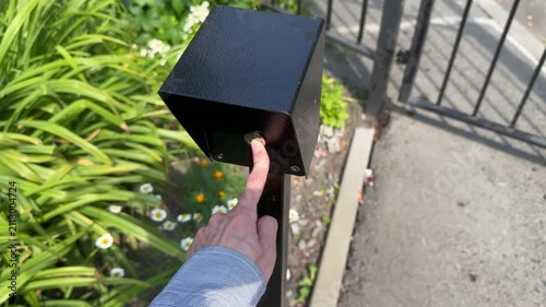 Hand reaches to press button on black box near gate, with green plants and flowers visible in the surrounding area