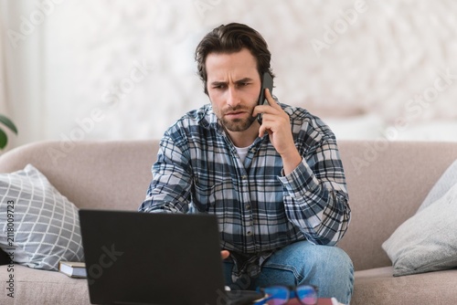 Foto Worried Man In His 30s, Caucasian, Sitting On Sofa While Using Laptop And Talking On Phone At Home, Engaged In Work