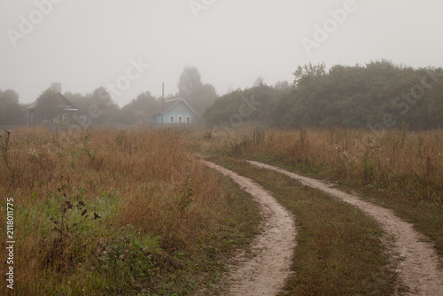 Dirt road through an autumn field leading to houses in morning fog