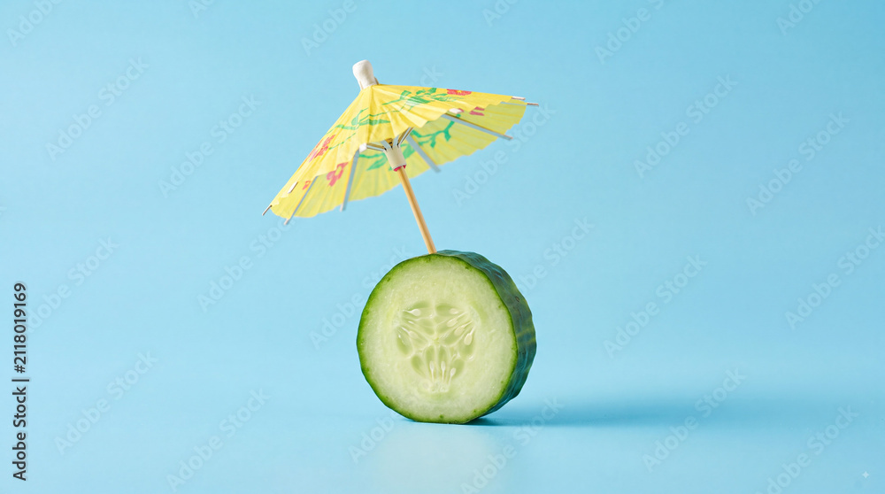 Naklejka premium Fresh green cucumber slice with a small yellow paper cocktail umbrella on a bright light blue background, creative food concept for summer and relaxation