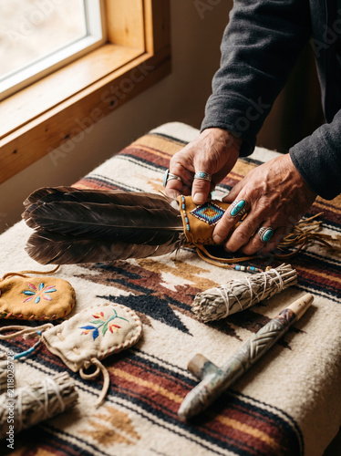 Hands arranging eagle feathers with turquoise rings on patterned textile near window, surrounded by sage bundles and decorated pouches, evoking calm and spiritual atmosphere