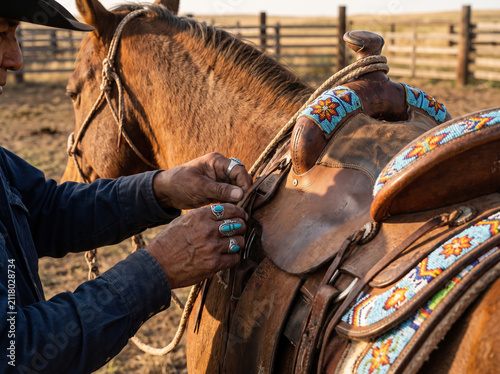 Man adjusting leather saddle on brown horse with colorful beaded decoration in outdoor corral during sunset