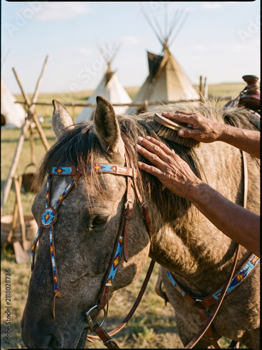 Hands grooming horse with beaded bridle near traditional teepee tents in outdoor camp setting on sunny day
