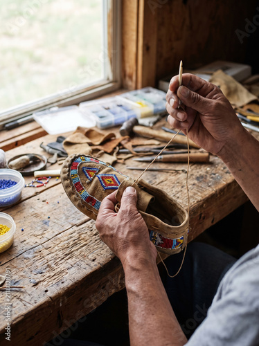 Hand sewing leather moccasin with colorful beadwork on wooden table near window, traditional craft with tools and beads, detailed artisan work creating cultural footwear
