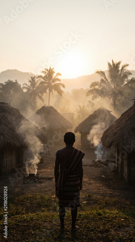 Tribe member standing outside traditional hut at sunrise with smoke rising and palm trees in background creating peaceful atmosphere
