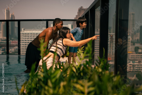 Family enjoying rooftop pool, woman pointing at urban city view together