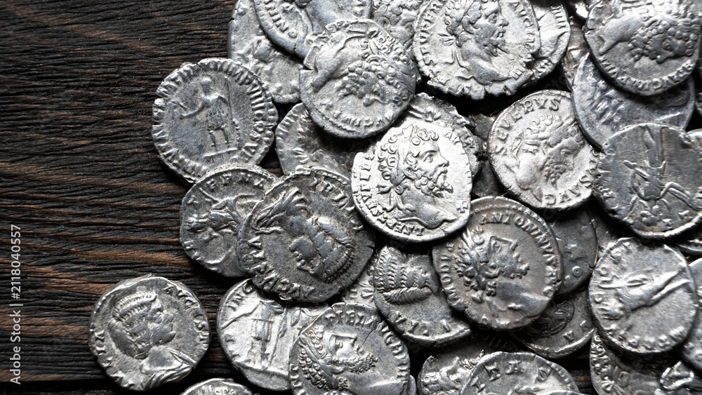 Obraz premium Pile of ancient Roman silver coins on a dark wooden background. Collection of silver denarii featuring portraits of emperors. Historical numismatics and archaeology concept.