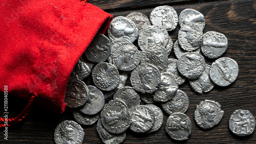 Ancient silver coins spilling from a red cloth pouch onto a dark wooden table. Roman denarii treasure hoard. Numismatics and historical wealth concept