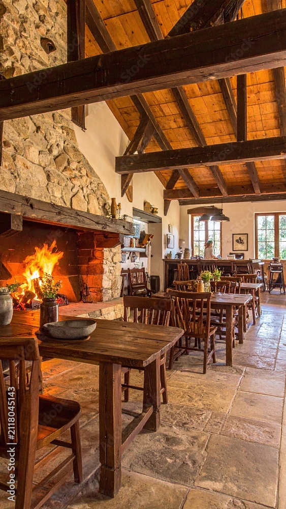 Obraz premium This cozy, rustic restaurant interior features a roaring stone fireplace, wooden furniture, and vaulted timber beams, evoking warmth, comfort, and tranquility with no people inside.