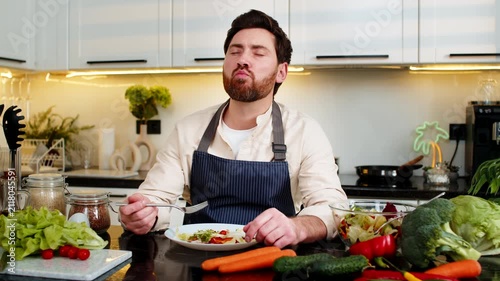 Middle-aged man enjoying fresh salad at home kitchen closing eyes in pleasure after healthy choice for diet. Young guy eating vegetarian lunch indoors using fork on plate feeling quite satisfied calm.