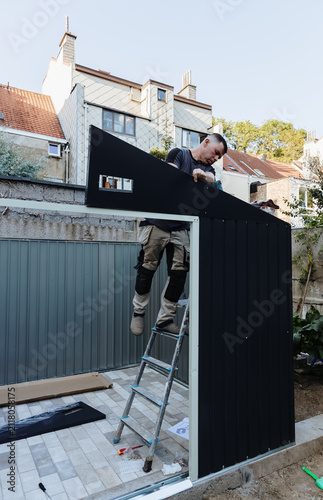 A handsome adult man with a drill installs roofing on a metal profile shed.