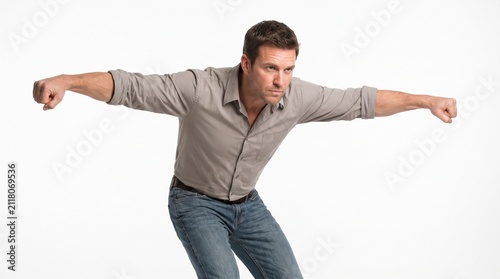 A determined man in a grey collared shirt and blue jeans leans forward with his arms outstretched, fists clenched, as if preparing for a powerful action or struggle.
