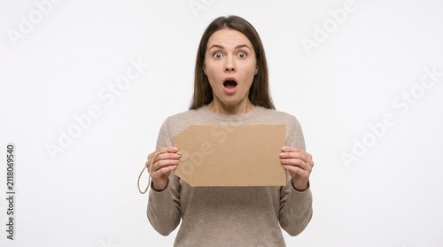 A surprised woman holding a blank tag, with her mouth agape in shock. She has her eyes wide open and is looking directly at the camera. Her expression conveys a sense of disbelief and astonishment