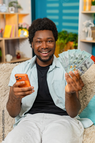 African American young man on home sofa counts money dollars cash and checks smartphone after online game win in living room. Black guy compares amount and enjoys sports bet or casino success indoors.