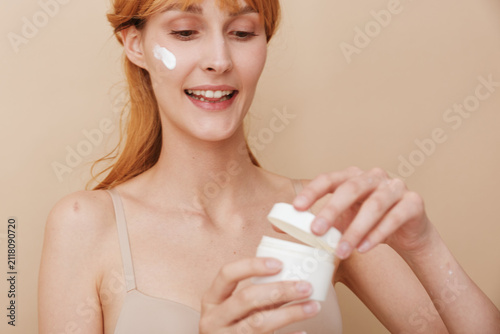 Photography A young adult woman with redhead hair applies moisturizer to her face, smiling softly in a beige indoor setting, showcasing a candid skincare moment of selfcare and calm