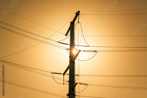 Obraz na plátně A power transmission tower stands as a silhouette against a warm sunset sky, with the sun partially hidden behind its structure