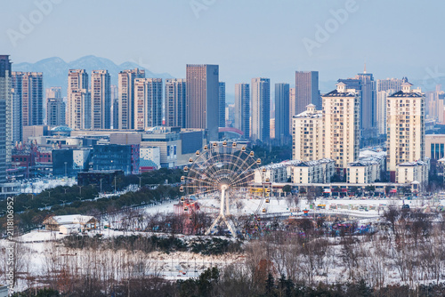 Konstfotografi A wintry urban landscape featuring a snow-covered park with a ferris wheel, surrounded by high-rise buildings, with distant mountains under a clear sky