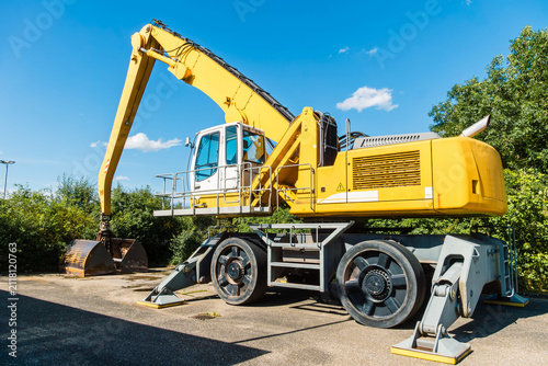 Yellow excavator with a grapple claw stands parked near greenery in Germany, heavy machinery equipment, industrial vehicle scene.