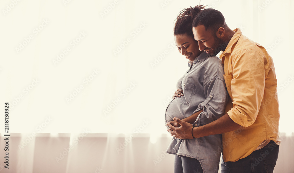 Fototapeta premium Joyful African American Couple Smiling While Embracing Pregnant Belly In Bright Room Filled With Natural Light. Happy Expecting Parents Enjoying Intimate Moment Together In Family Setting.